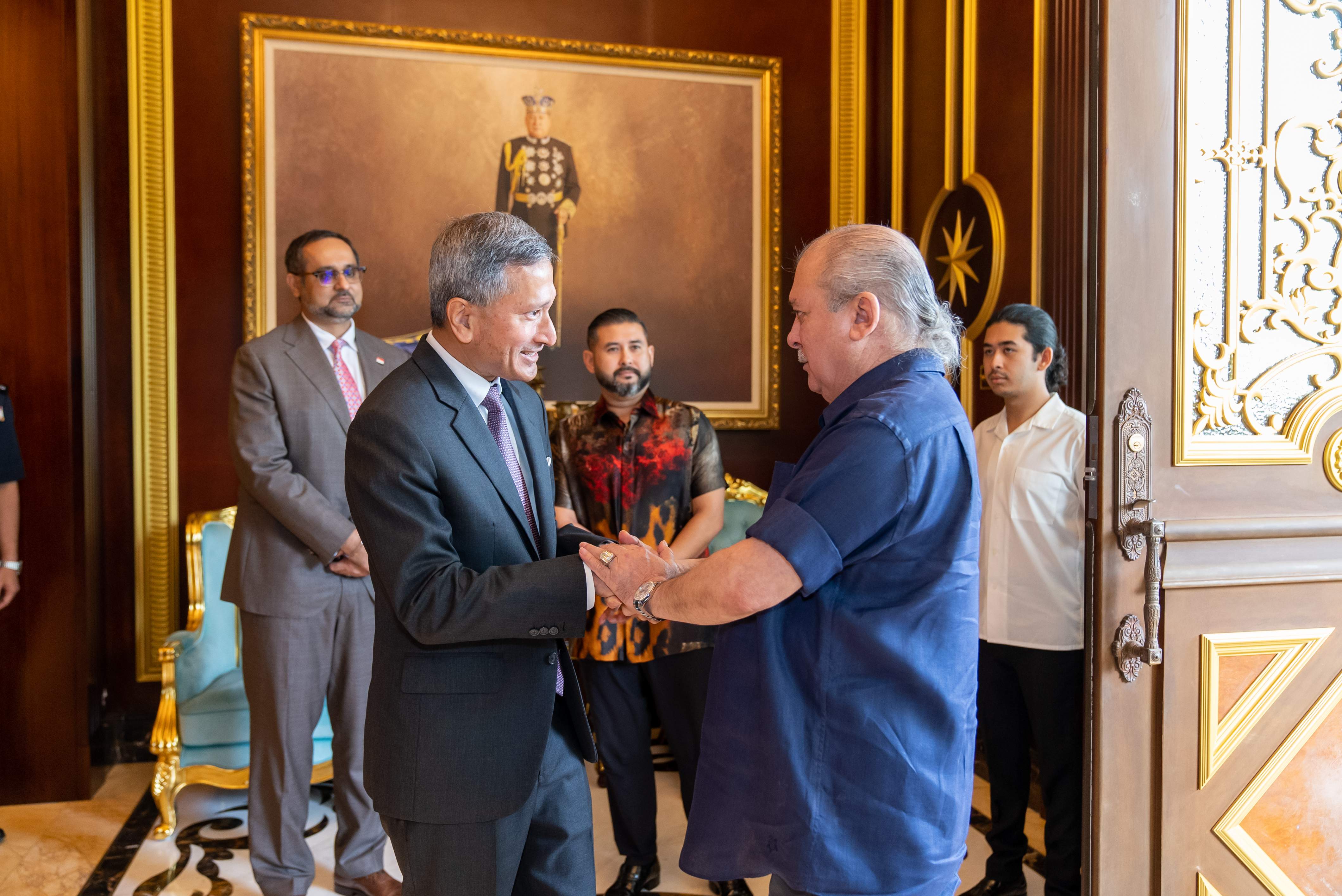 Two men in suits shaking hands, watched by three other men in front of a portrait.